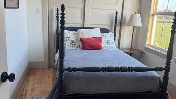 A bedroom with a four-poster bed, a patterned rug, a window with a view of greenery, and a door with a light switch.