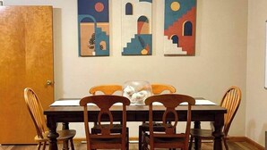 Een eethoek met een houten tafel en vier stoelen, een vaas met bloemen, en drie abstracte schilderijen aan de muur.