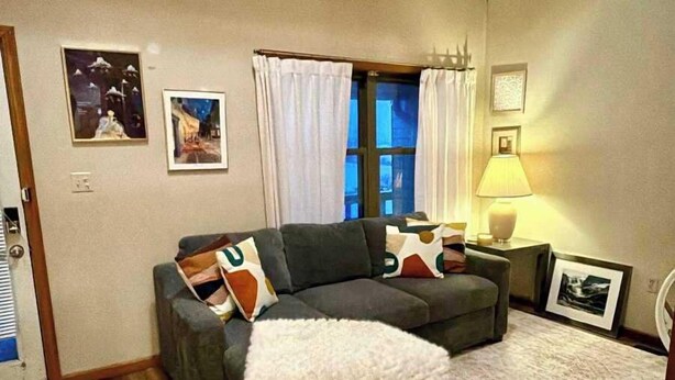 Een woonkamer met een hoekbank, een zacht, wit vloerkleed en een raam met gordijnen.