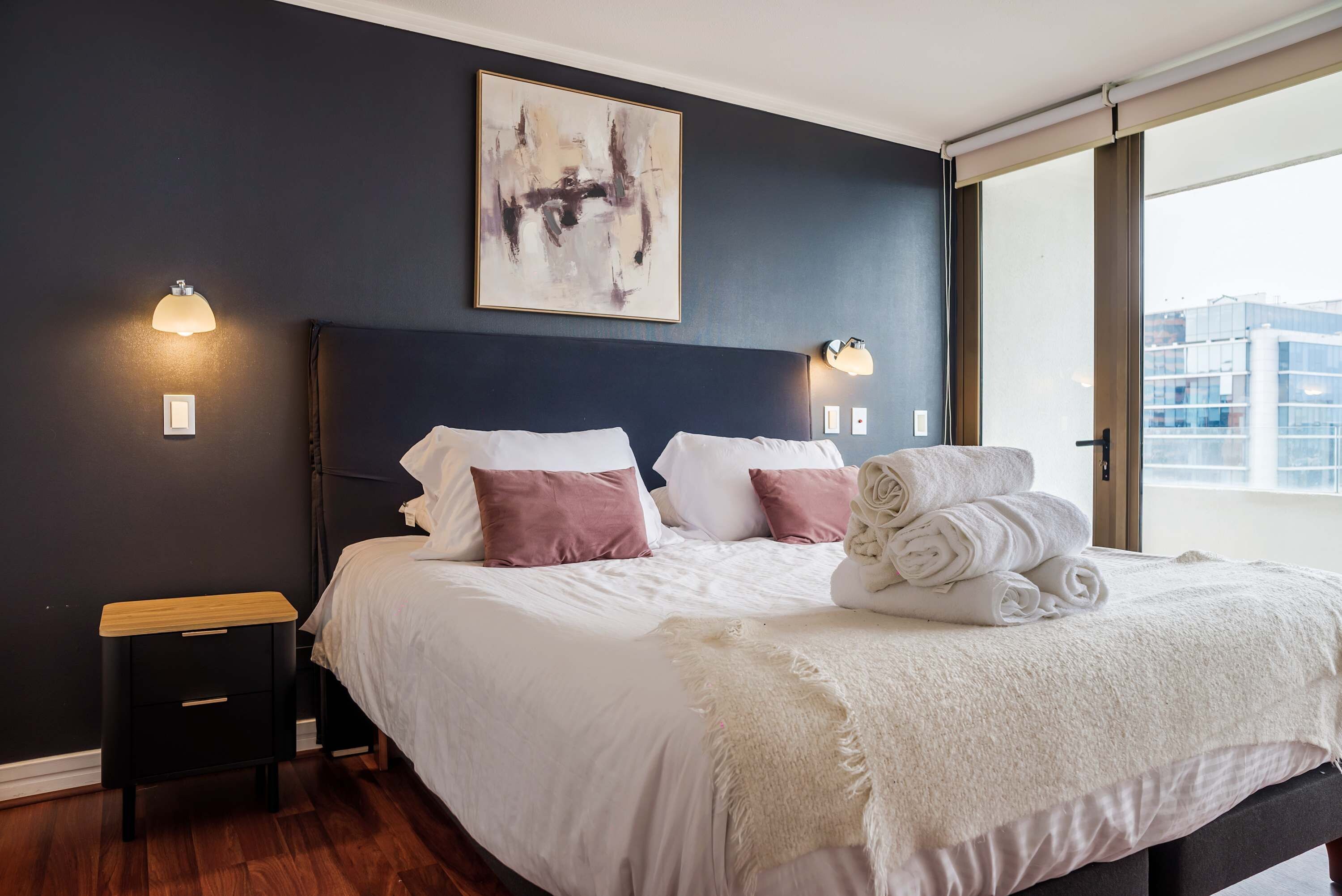 Une chambre moderne avec un grand lit, une table de chevet, une lampe fixée au mur et une fenêtre donnant sur la ville.