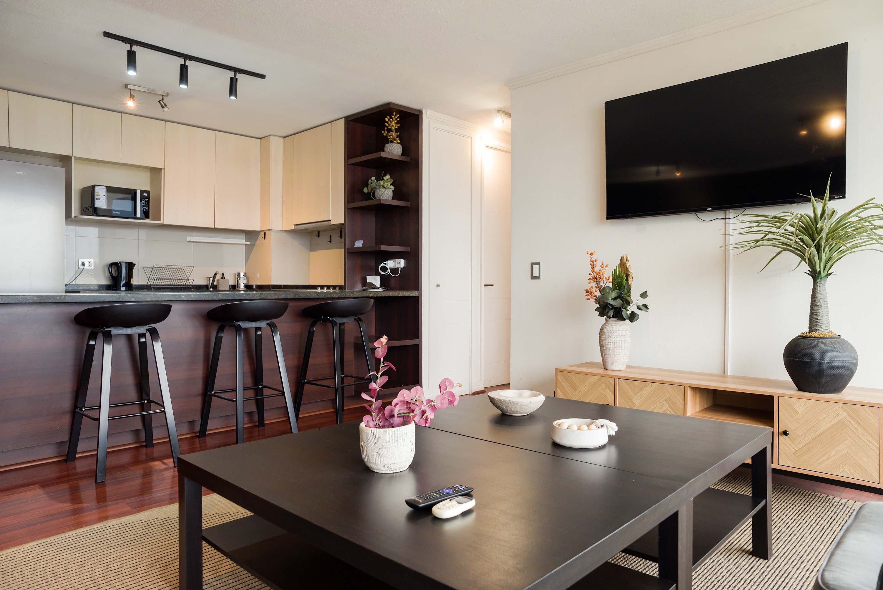 A modern kitchen with a microwave, a coffee machine, and a sink. There are three bar stools and a dining table with a vase of flowers. A flat-screen TV is mounted on the wall.