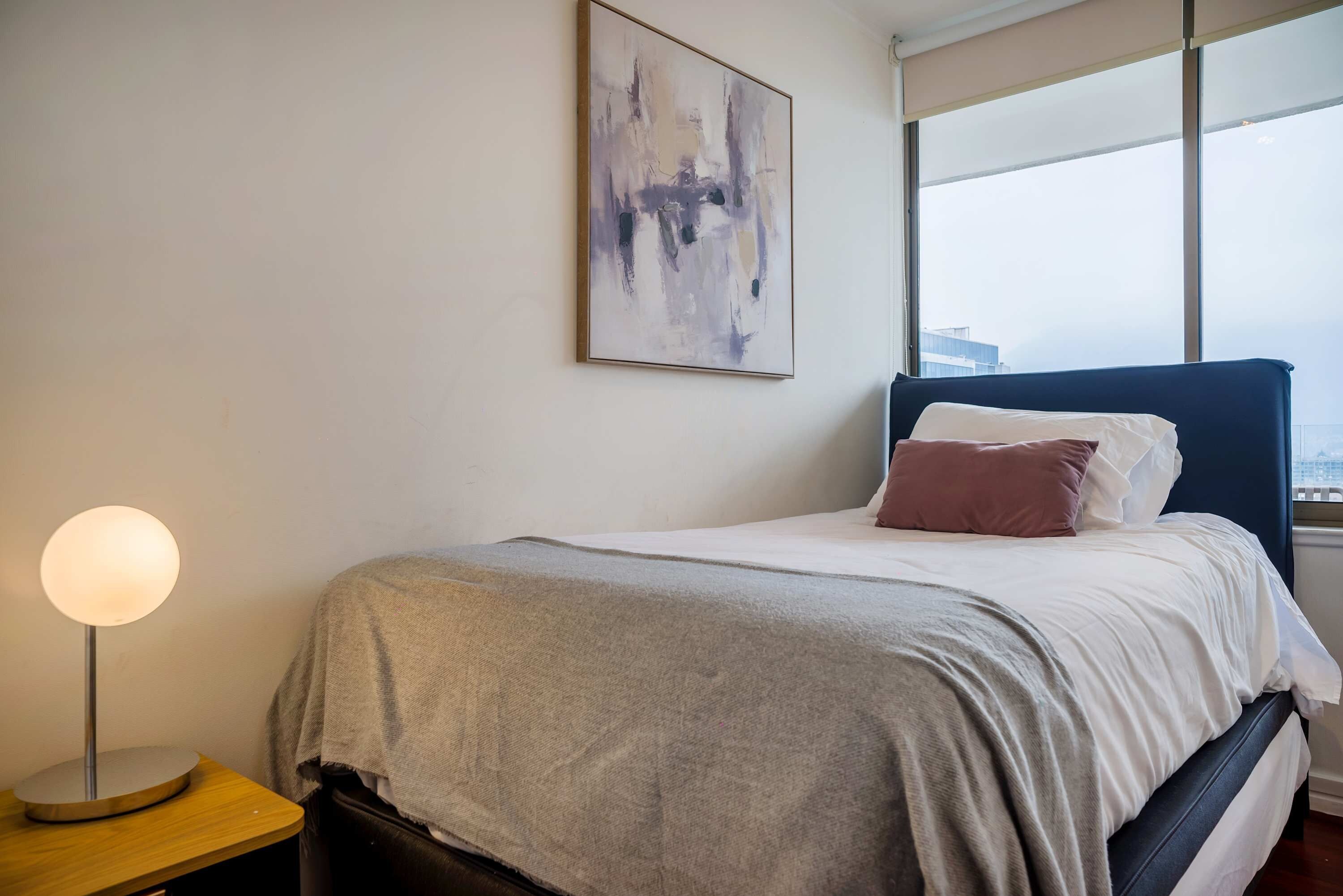Quarto de hotel com cama, abajur, janela com vista e pintura abstrata na parede.