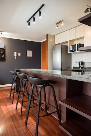 A modern kitchen with a bar area, wooden flooring, and stainless steel appliances. - Spacious 2 Bedroom Apartment with Pool and Gym! (Las Condes)