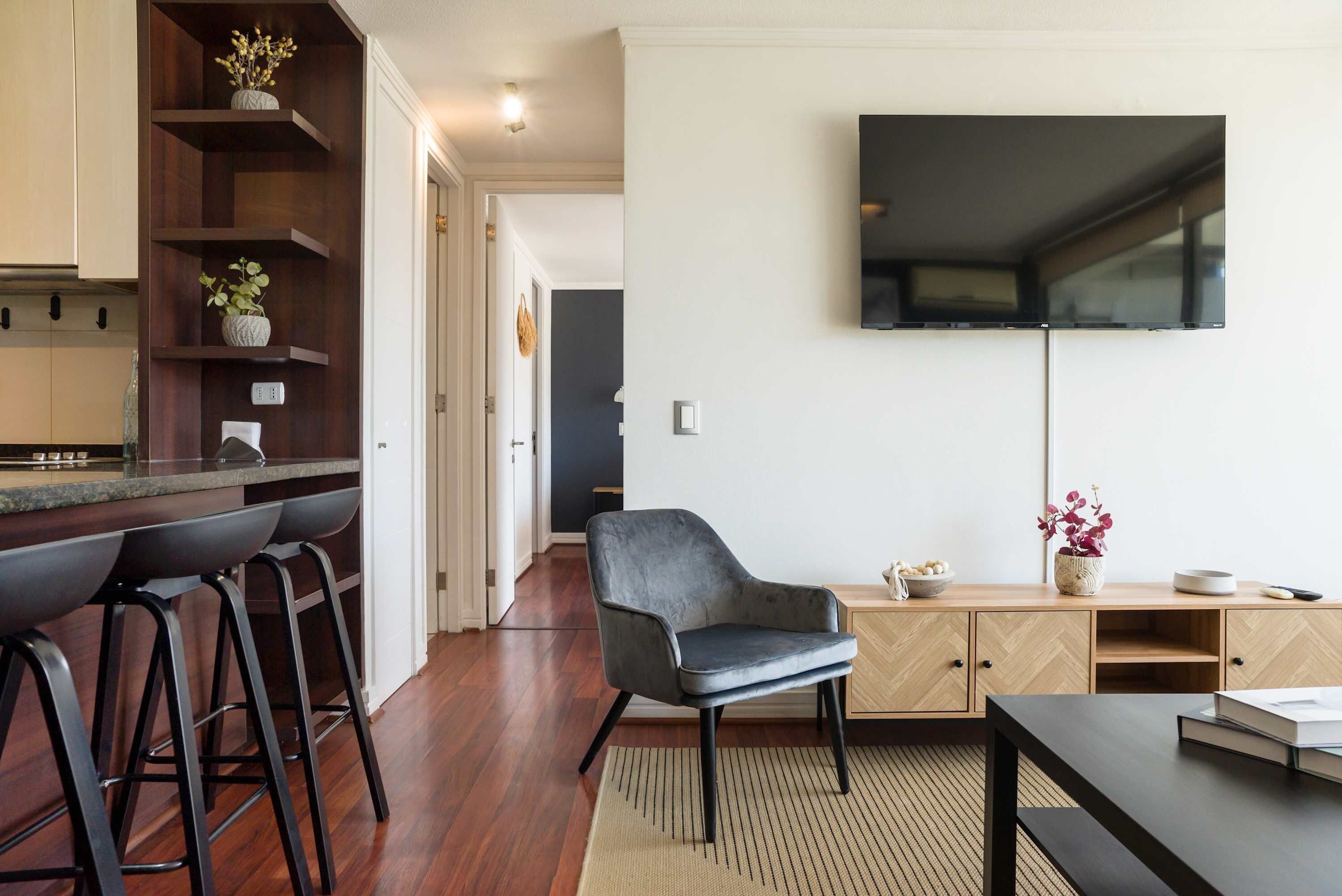 Private living room with a wall-mounted TV, gray lounge chair, woven rug, and open-plan kitchen with bar seating.