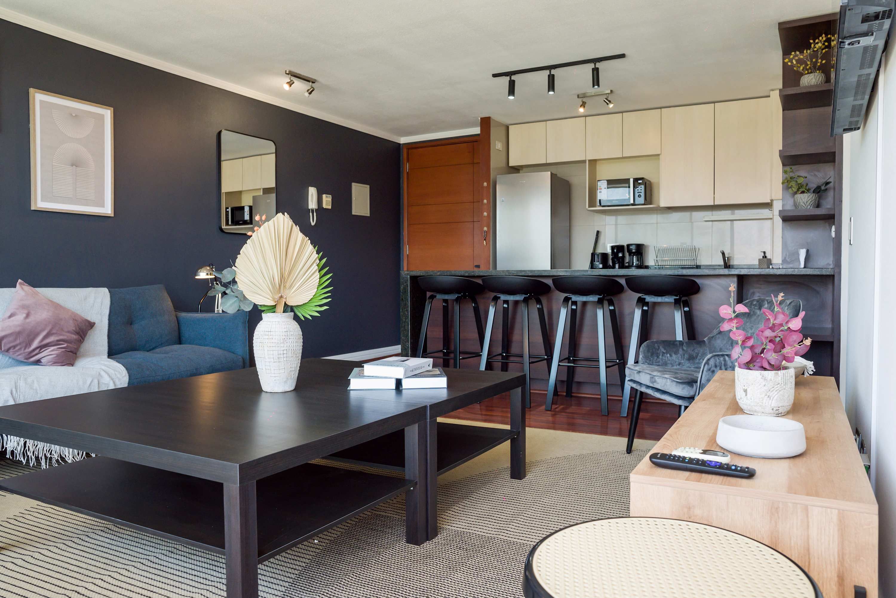 Una sala de estar moderna con un sofá oscuro, una mesa de centro y una cocina con microondas y taburetes de bar.