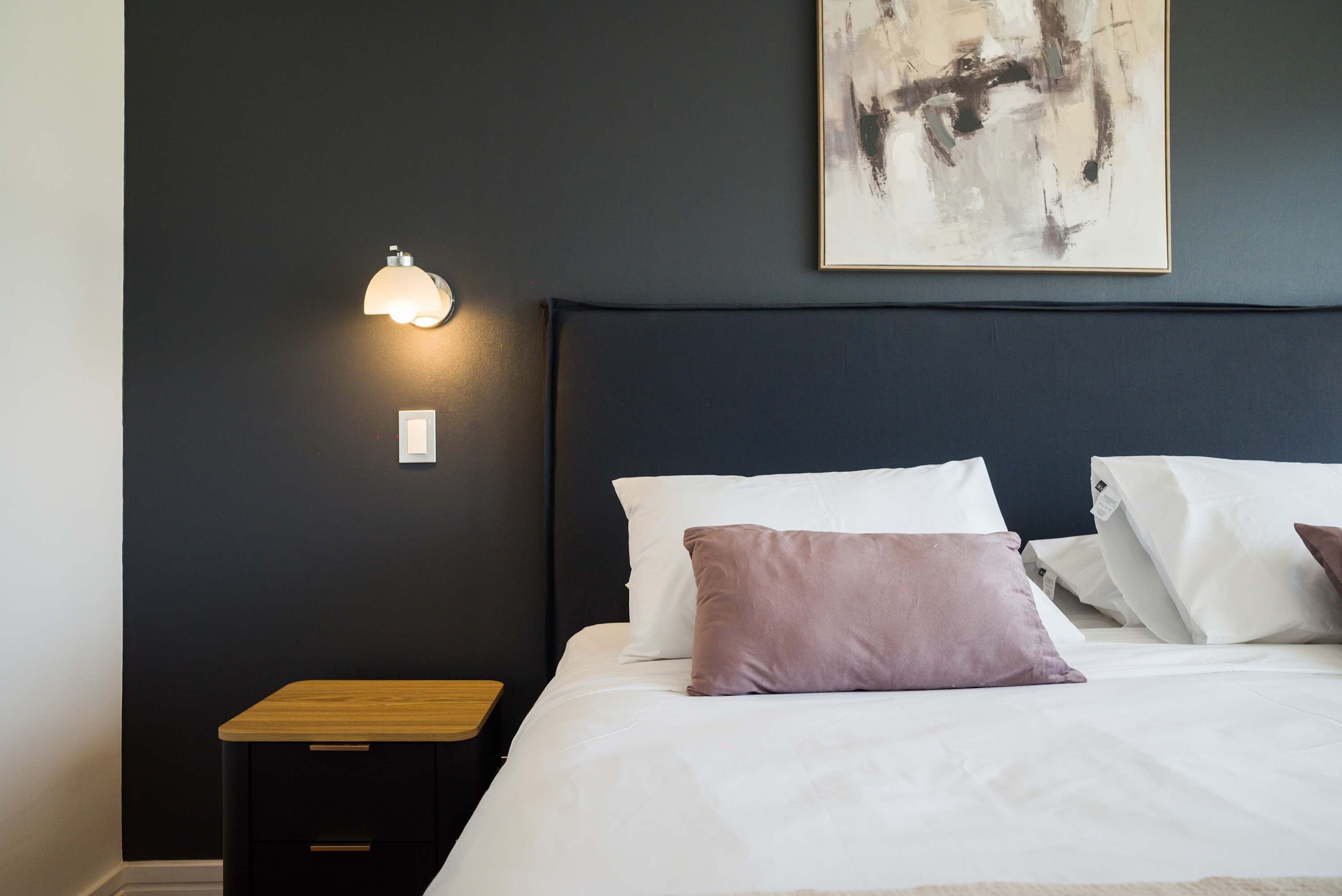 A bedroom with a dark wall, a bedside table, a lamp, a bed with white bedding and a single purple pillow, and a piece of abstract art above the bed.