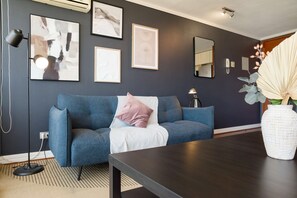 Un salon moderne avec un canapé bleu foncé, un plaid blanc et une table basse en bois foncé. On peut voir des œuvres d’art encadrées au mur, un lampadaire et un vase blanc rempli de fleurs.