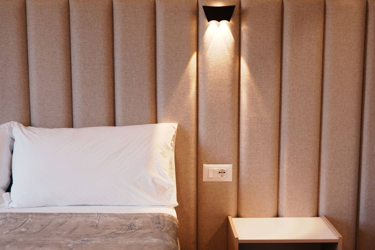 Una cama bien hecha con una almohada y una toalla enrollada, una mesita de noche con cajón, y una lámpara de pared.