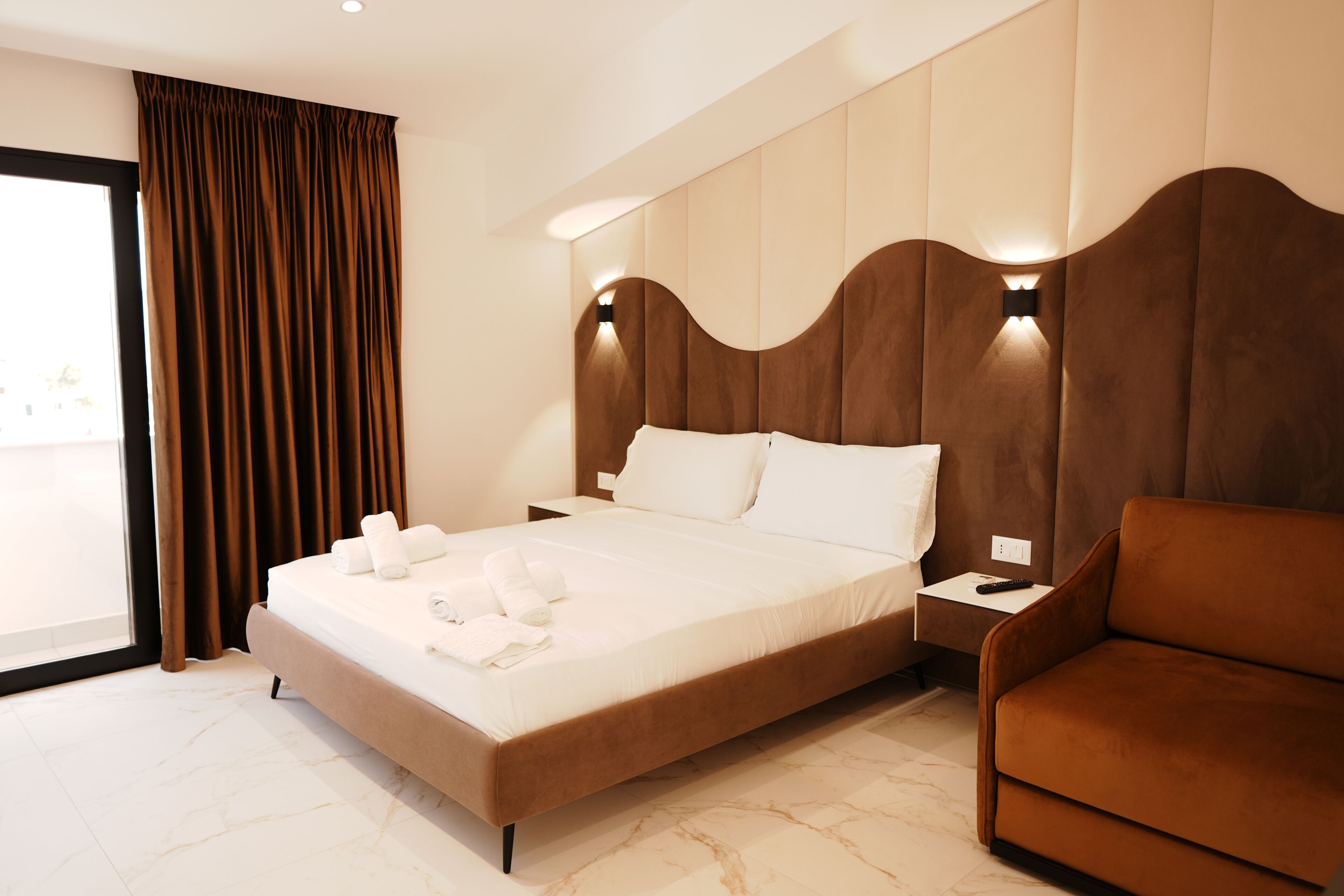 Une chambre d’hôtel moderne avec un grand lit, un fauteuil brun et une table de chevet avec une télécommande.