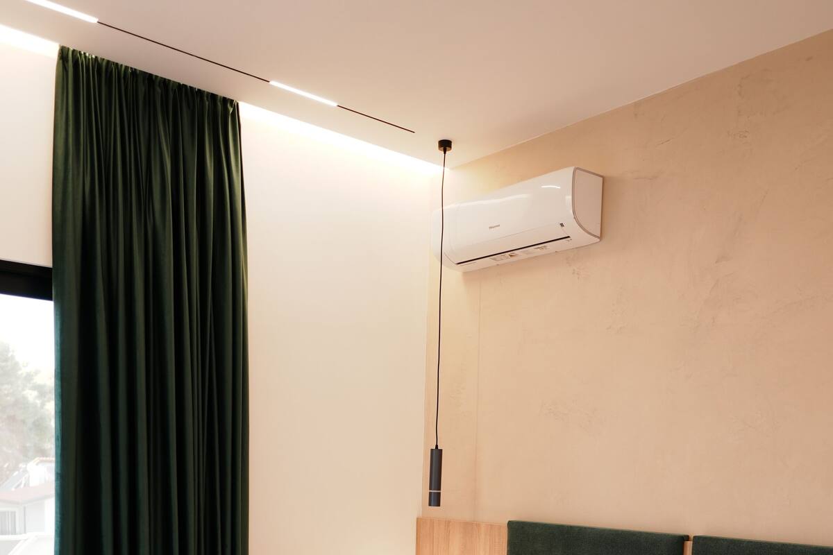 Una cama bien hecha con ropa y toallas blancas, un cabecero de madera, una mesita de noche con una lámpara y un equipo de aire acondicionado en la pared.