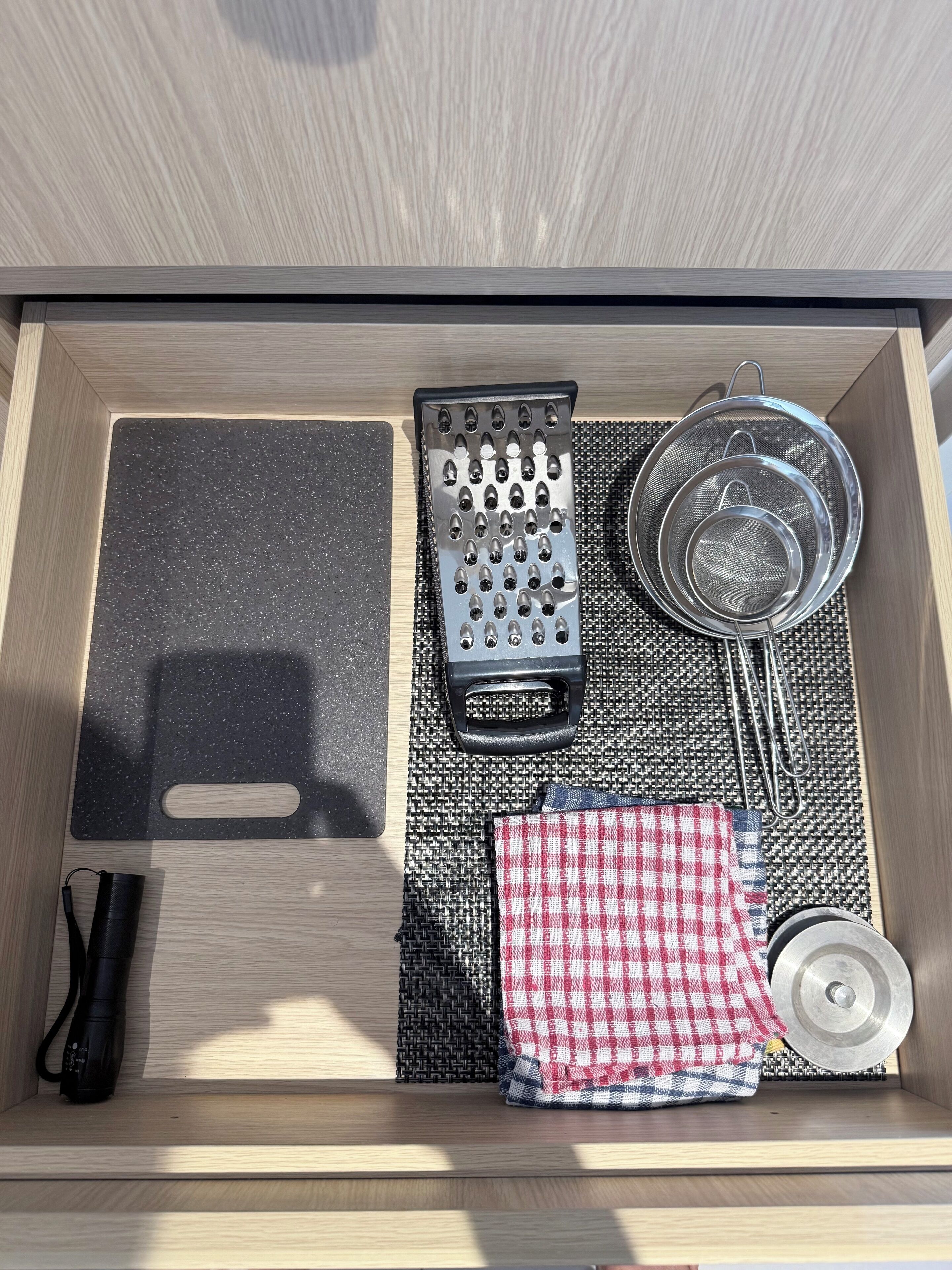 A drawer with a black cutting board, a grater, a metal strainer, a checkered cloth, a metal bowl, and a black flashlight.