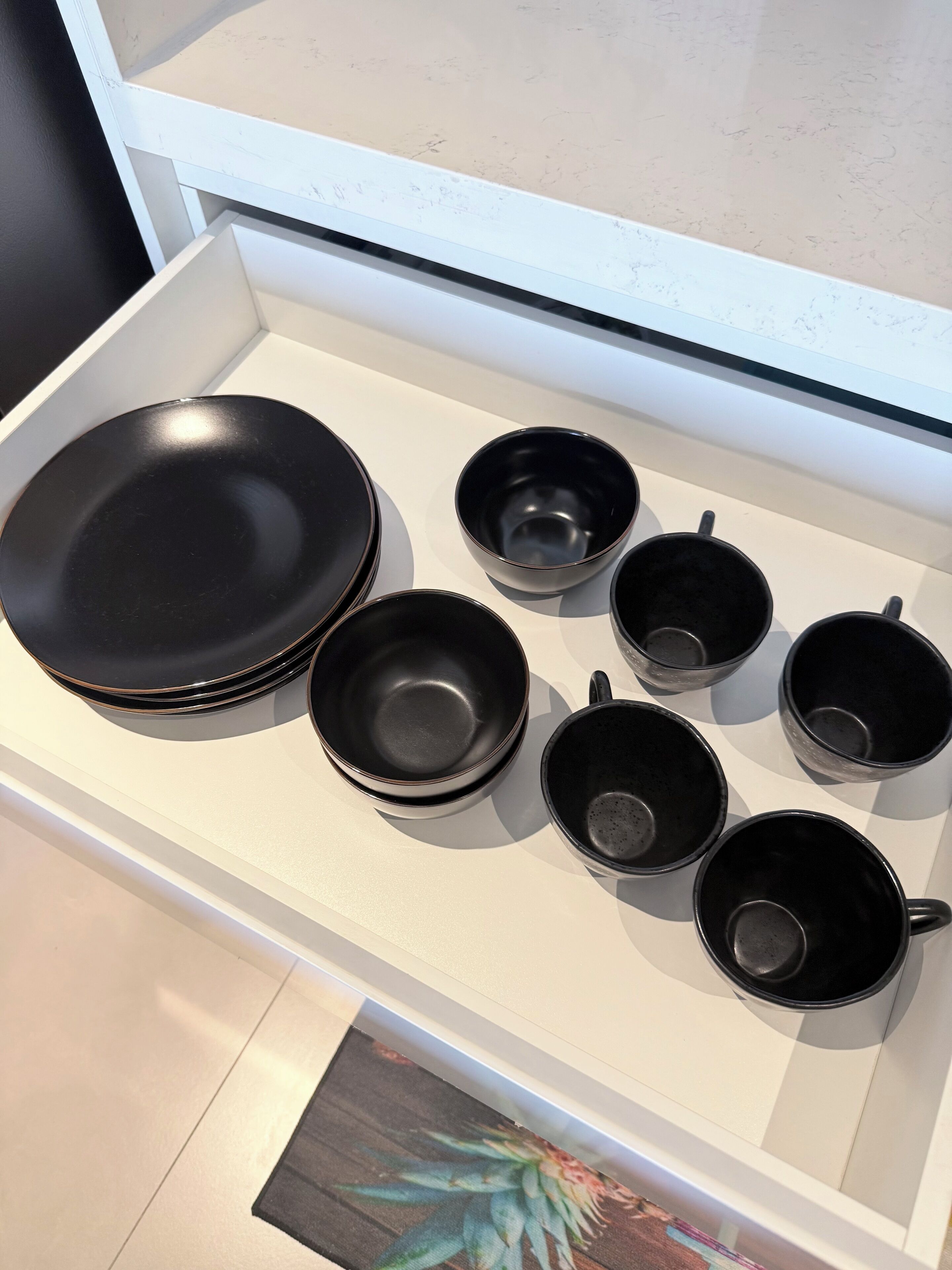 Une collection d’assiettes et de tasses en céramique noire est exposée sur une étagère blanche.