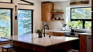 A modern kitchen with a large island, wooden cabinets, and a view of a forest through large windows. - The Alpenglow: Waterfront Escape Private Hot Tub (Juneau)