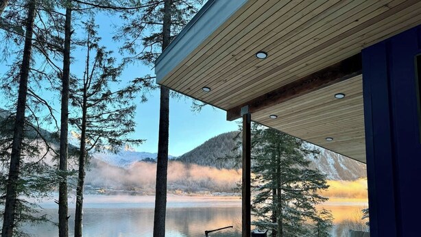 A cabin with a wooden deck overlooking a lake, surrounded by trees and mountains. - The Alpenglow: Waterfront Escape Private Hot Tub (Juneau)
