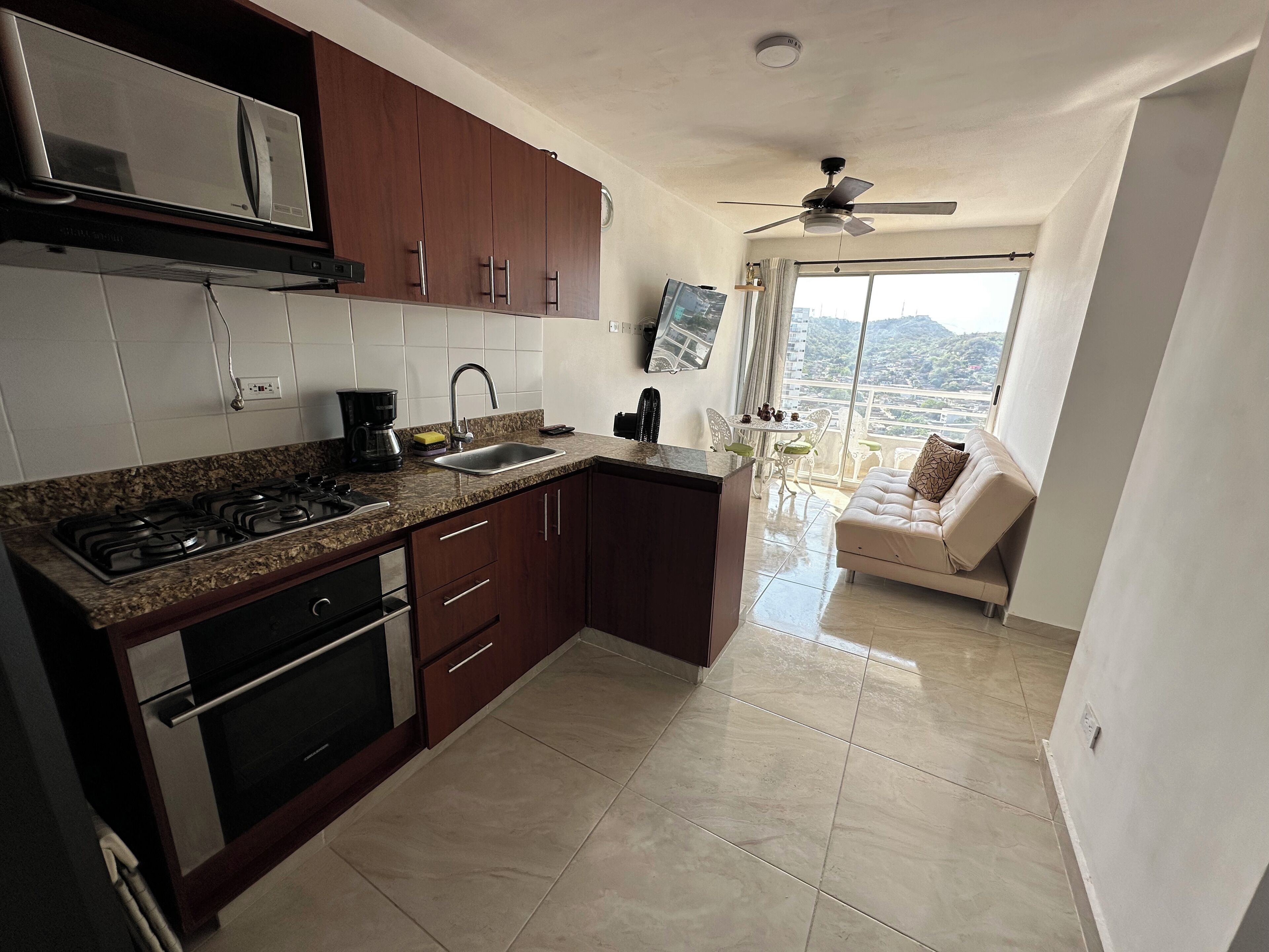 Cozinha moderna com bancadas de granito, eletrodomésticos de aço inoxidável e armários de madeira. Há uma área de jantar com sofá e vista da cidade através de grandes janelas.