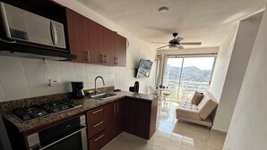A modern kitchen with granite countertops, stainless steel appliances, and wooden cabinets. There is a dining area with a sofa and a view of the city through large windows. - Edificio Lotus - Torices (Cartagena)