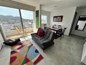 A living room with a grey sofa, a colorful rug, a dining table with chairs, a balcony with a chair, and a city view. - Edificio Lotus - Torices (Cartagena)