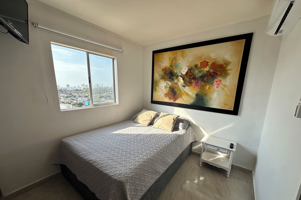 Quarto com cama, janela vista para a cidade, um quadro e mesa de cabeceira.