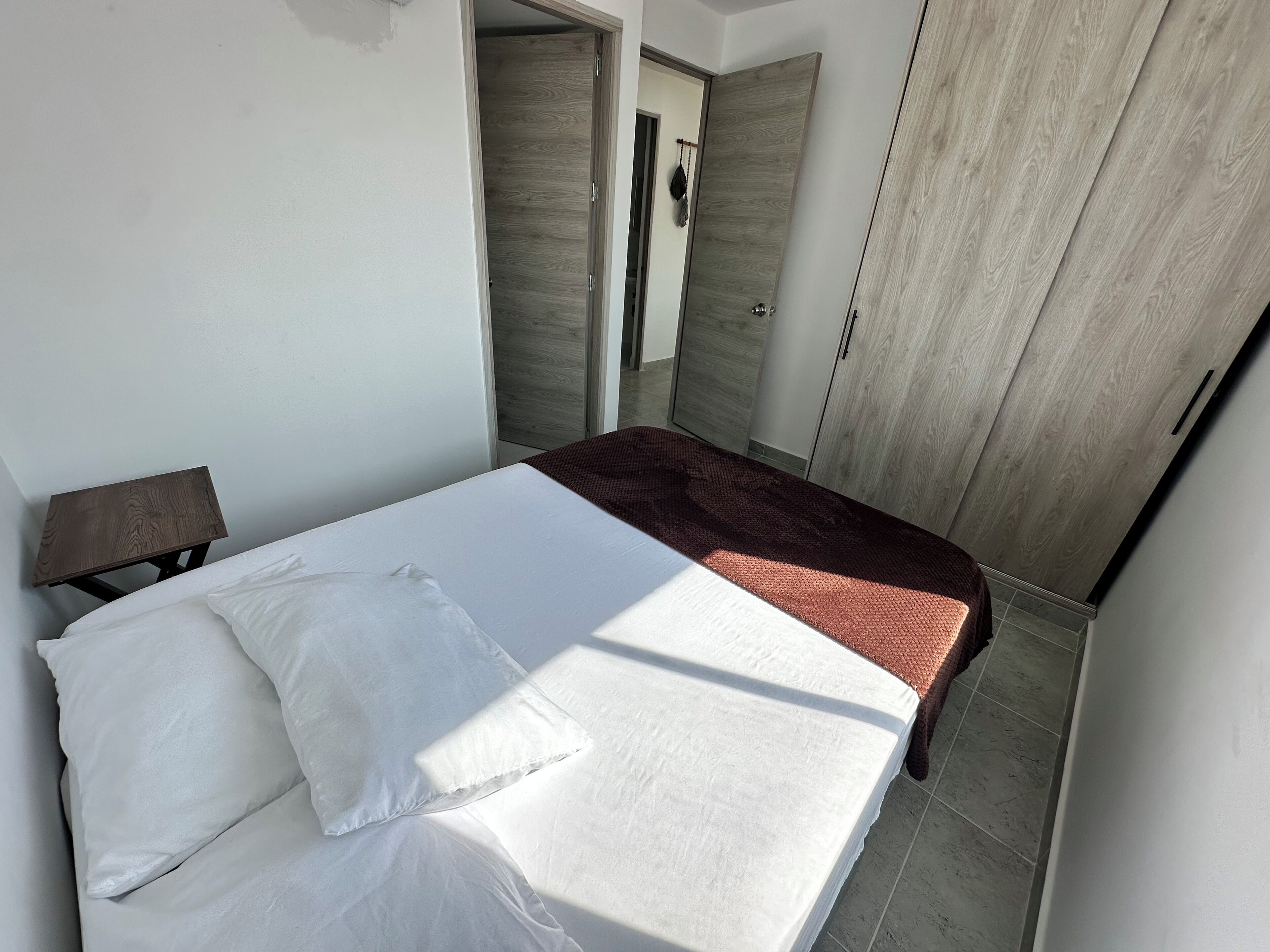 A bedroom with a bed, pillows, a wooden nightstand, a red throw blanket, and a wardrobe with a mirror.