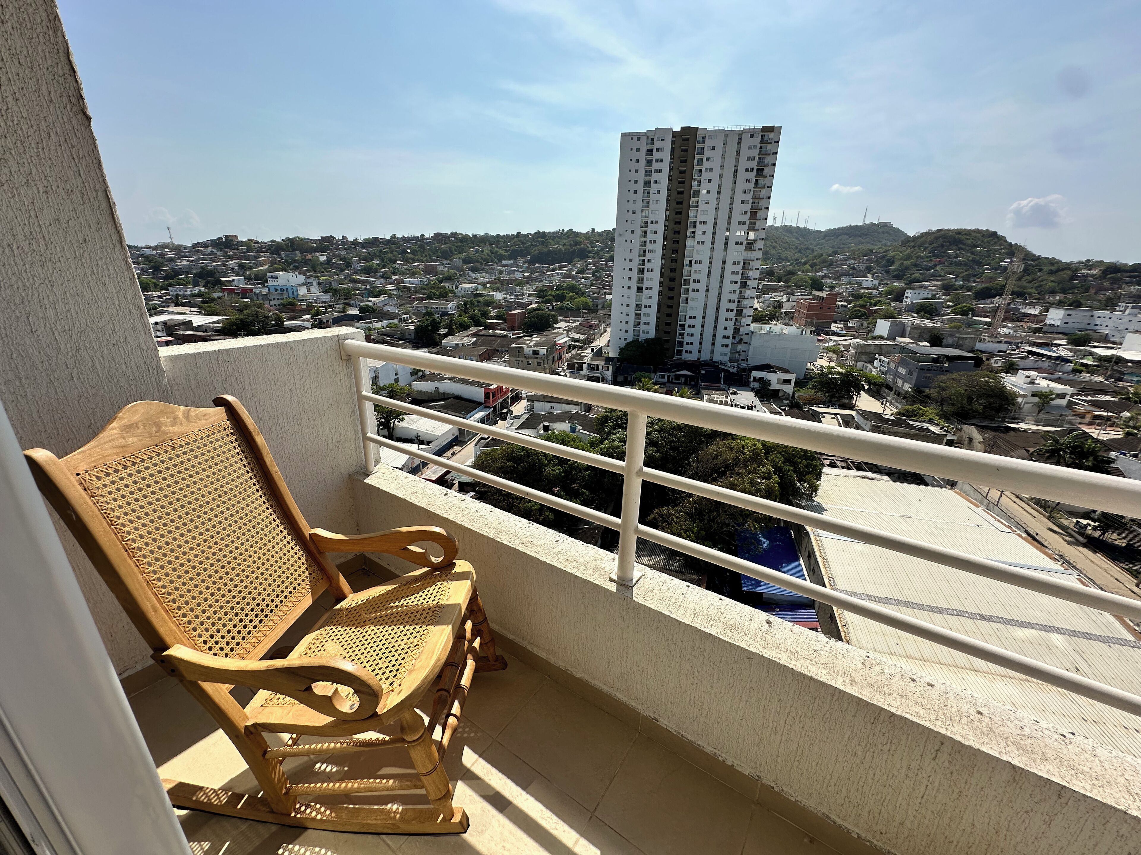 A balcony with a wooden chair, a white railing, and a view of a cityscape including a tall building.