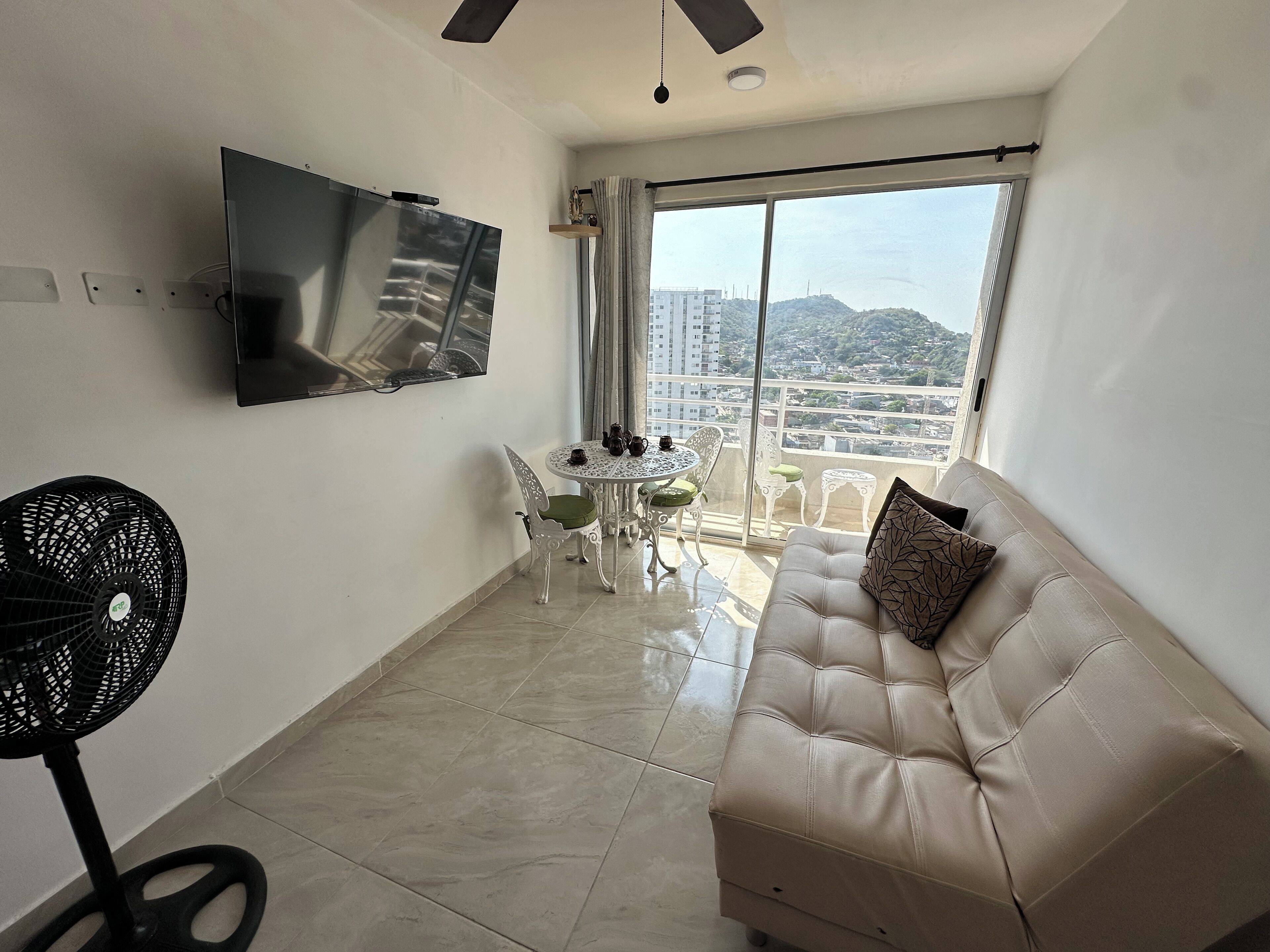 A modern living room with a beige sofa, a TV mounted on the wall, a dining table with chairs, and a large window offering a city view.