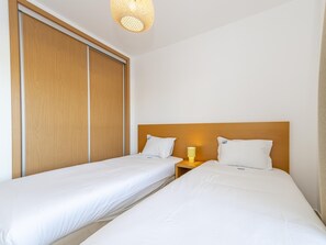 A hotel room with two single beds, a wooden wardrobe, and a bedside table with a lamp. - Beachside Bliss 5 mins Beach & Waterfront - Pool - Wi-Fi (Cabanas de Tavira)