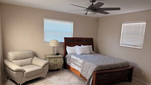 Une chambre à coucher comprenant un lit, une chaise, une table de chevet, un ventilateur de plafond et deux fenêtres équipées de stores.