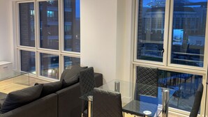 Una sala moderna con una mesa de comedor de vidrio y sillas, un sofá de color oscuro y ventanas amplias que ofrecen vistas panorámicas de la ciudad.