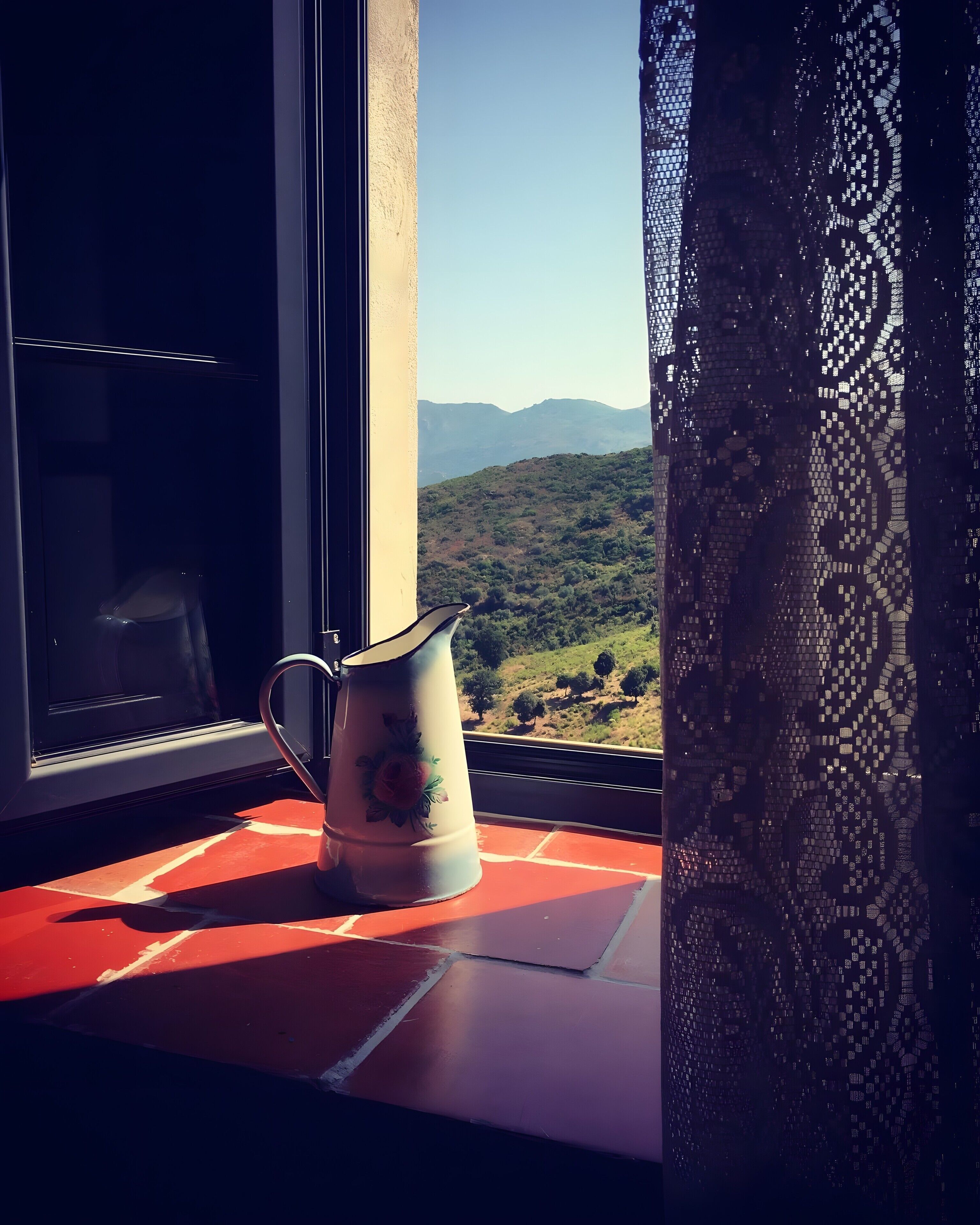 A window with a view of a mountainous landscape, a decorative pitcher on a red surface, and a patterned curtain.