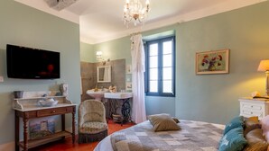 Une chambre à coucher comprenant un lit, une chaise, une coiffeuse, un téléviseur et une fenêtre avec des rideaux.
