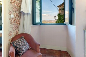 A corner room with a view of a mountain landscape through a window, a chair with a cushion, and a curtain with a floral pattern. - Casa Ghjunca Guesthouse – Ciucciarella Room (Rapale)