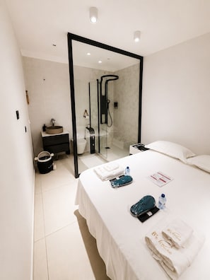 A modern hotel room with a bed, a bathroom with a sink and shower, and a large mirror.