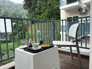 A balcony with a white chair, a tray with a cup, a bottle, and a plate of food. - Hotel Abline The Oaks (Rishikesh)