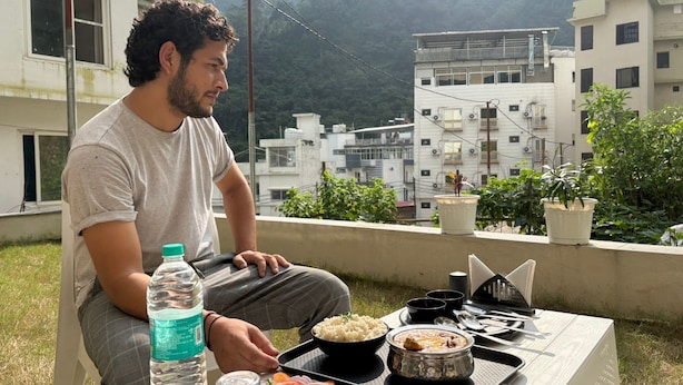 A person sitting at a table with a meal, a water bottle, and a view of buildings and greenery. - Hotel Abline The Oaks (Rishikesh)