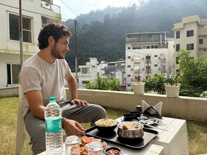 A person sitting at a table with a meal, a water bottle, and a view of buildings and greenery. - Hotel Abline The Oaks (Rishikesh)