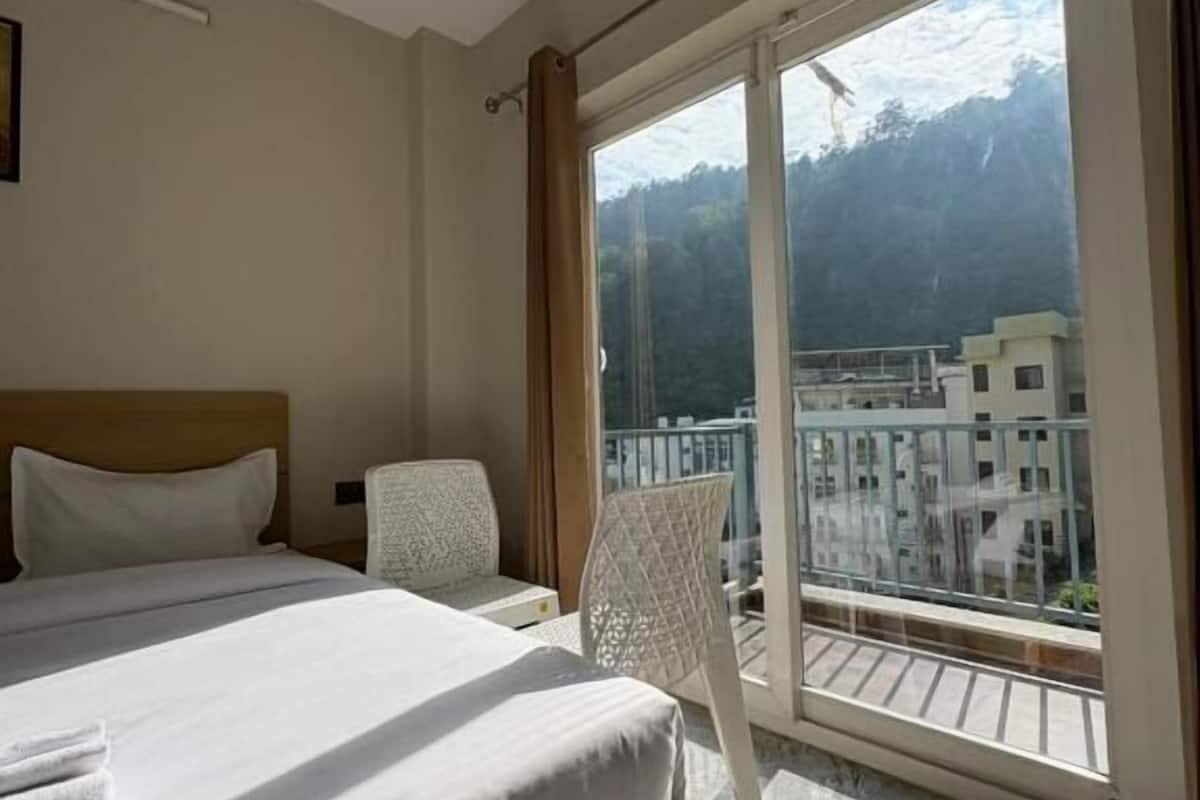 Habitación de hotel con una cama grande, una silla y un balcón con vistas a edificios y montañas.