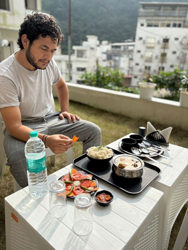 A person sitting at an outdoor table with a meal, a water bottle, and a tray with food and utensils. - Hotel Abline The Oaks (Rishikesh)