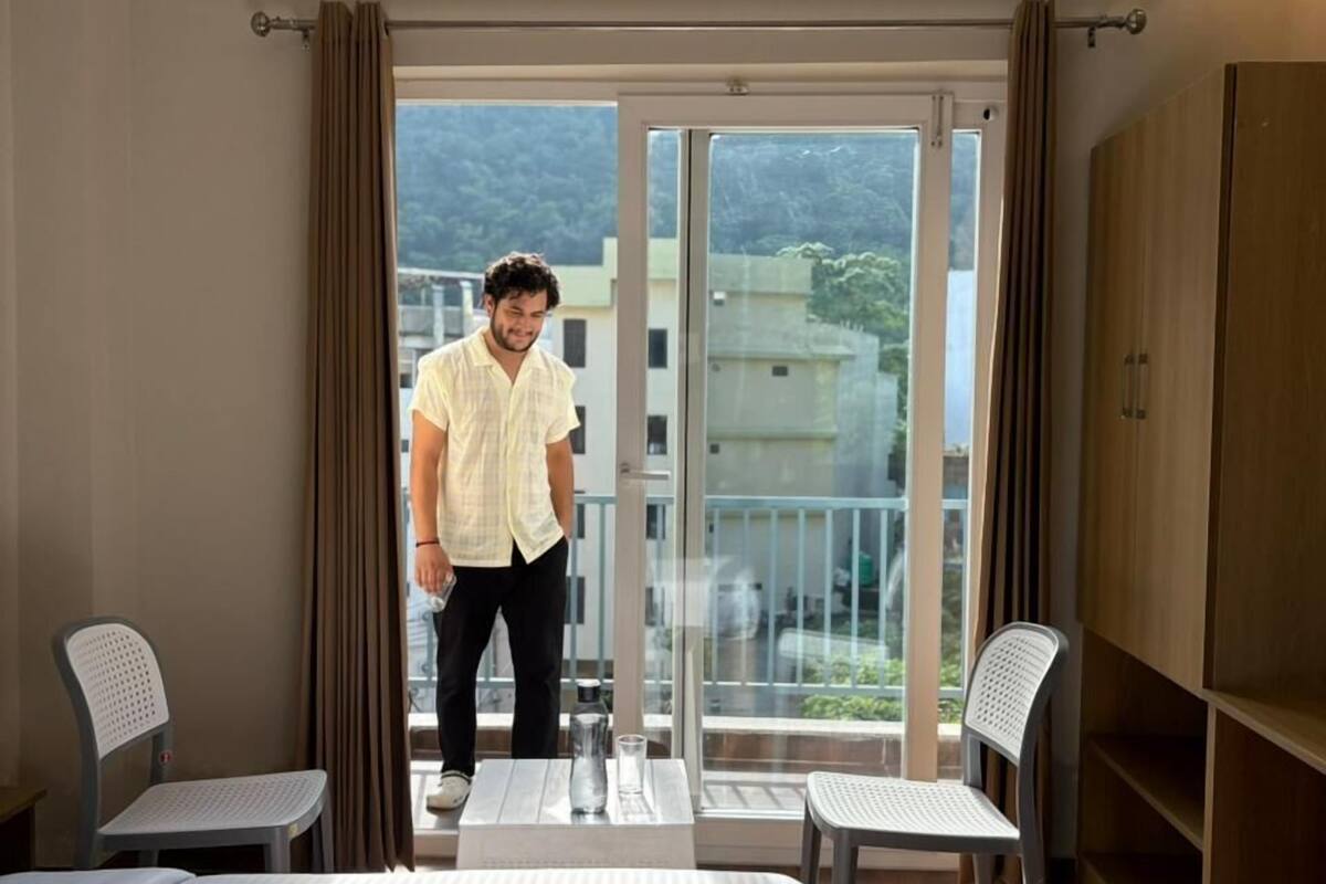 Habitación de hotel con cama, dos sillas, una mesa, armario y balcón con vista a edificios y árboles.