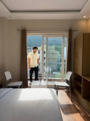 A hotel room with a bed, two chairs, a table, a wardrobe, and a balcony with a view of buildings and trees. - Hotel Abline The Oaks (Rishikesh)