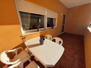 Une petite terrasse avec une table et des chaises blanches, une fenêtre avec des stores et des plantes en pot.