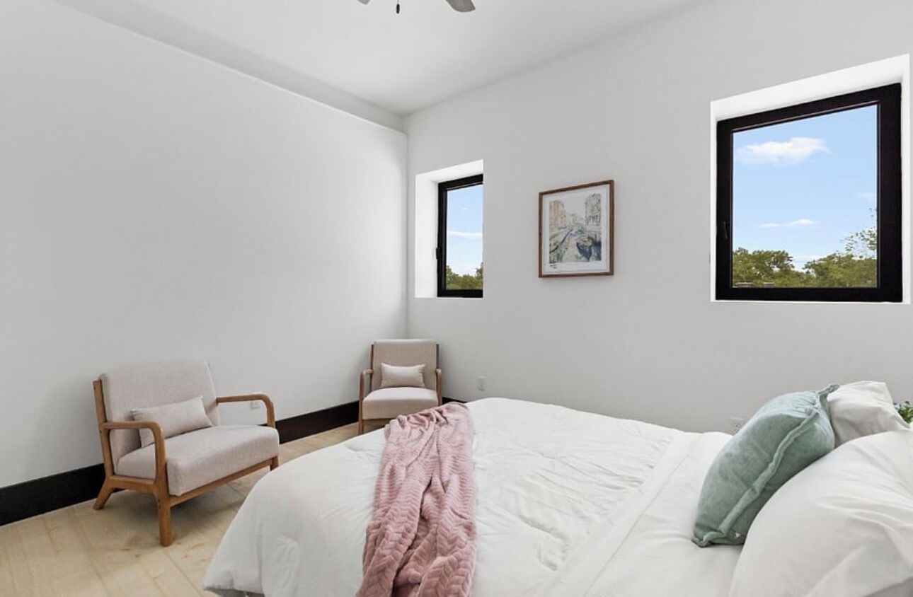 A modern bedroom with a large bed, two chairs, and two windows providing natural light.