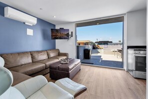 Un salon moderne avec un canapé en cuir marron, un divan en cuir blanc et un petit pouf. Il y a également un coin cuisine avec une cuisinière et un four à micro-ondes. Une grande fenêtre offre une vue sur l’extérieur.