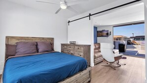 Une chambre à coucher avec un lit, une tête de lit en bois, un ventilateur de plafond, une commode, une chaise blanche et une vue sur l’extérieur.