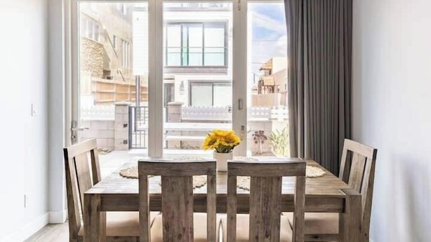 Uma sala de jantar com mesa e cadeiras de madeira, um vaso com flores amarelas e vista para uma varanda através de grandes portas de vidro.
