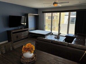 A modern living room with a flat-screen TV, a sofa, a coffee table, and a dining table with a vase of flowers.