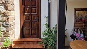 Uma porta de madeira com o número 5, uma parede de pedra, uma planta em vaso e uma cadeira com almofada colorida.