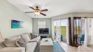A living room with a ceiling fan, a television, a sofa, and a balcony with curtains. - Walk to Beach, Dining & Shops- Community Pool (Holmes Beach)
