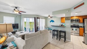 A modern living room with a sofa, TV, and a balcony with curtains. - Walk to Beach, Dining & Shops- Community Pool (Holmes Beach)