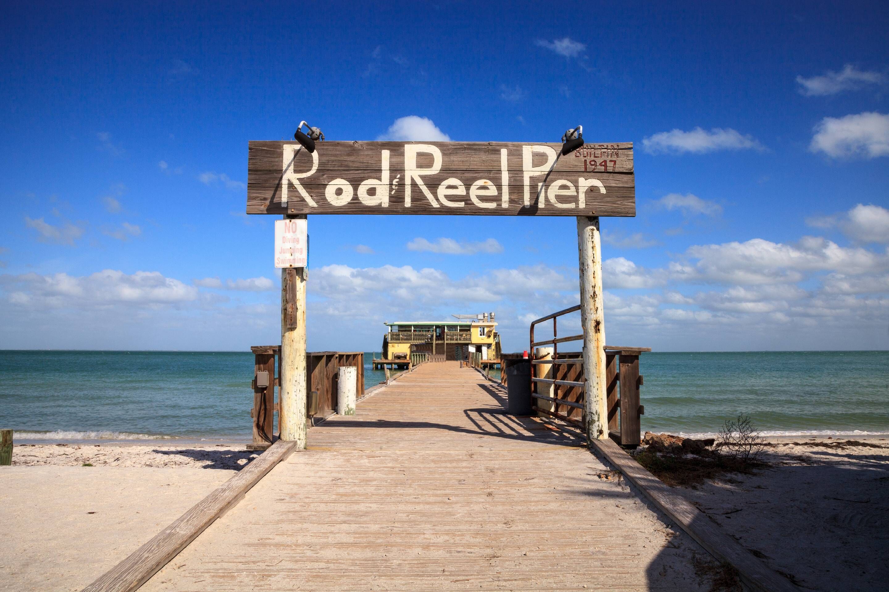 Een houten steiger die naar Rod Reeler leidt, met een helder uitzicht over de oceaan en een gebouw in de verte.