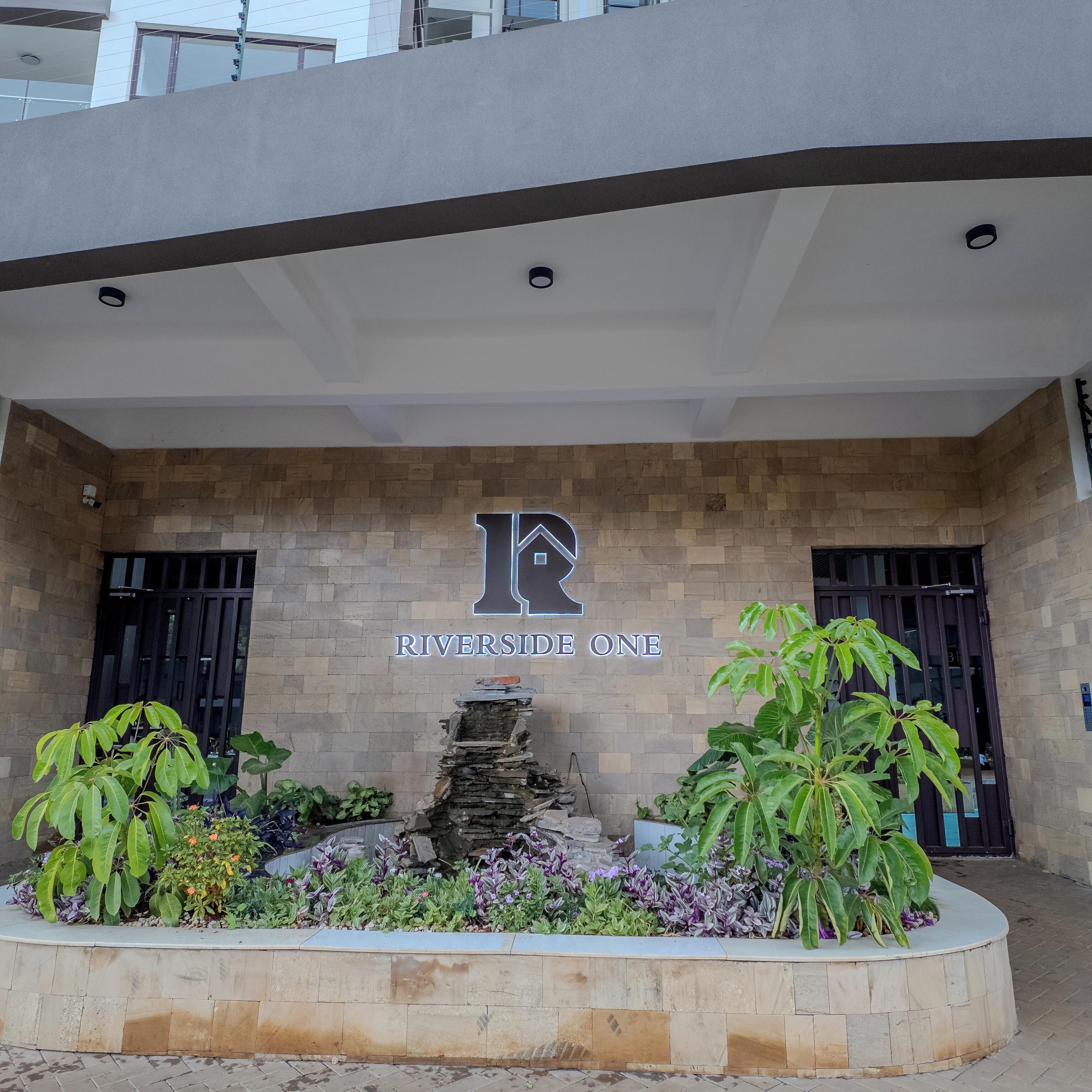 A building entrance with a sign reading "RIVERSIDE ONE" and a stone wall with plants.